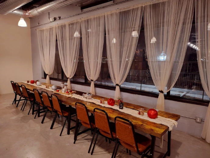 Modern event space with exposed ceiling and concrete floor. Wooden tables are placed in a long communal setup, decorated with a linen table runner and small trinkets. Image background shows tall windows draped with sheer curtains. It is nighttime outside.